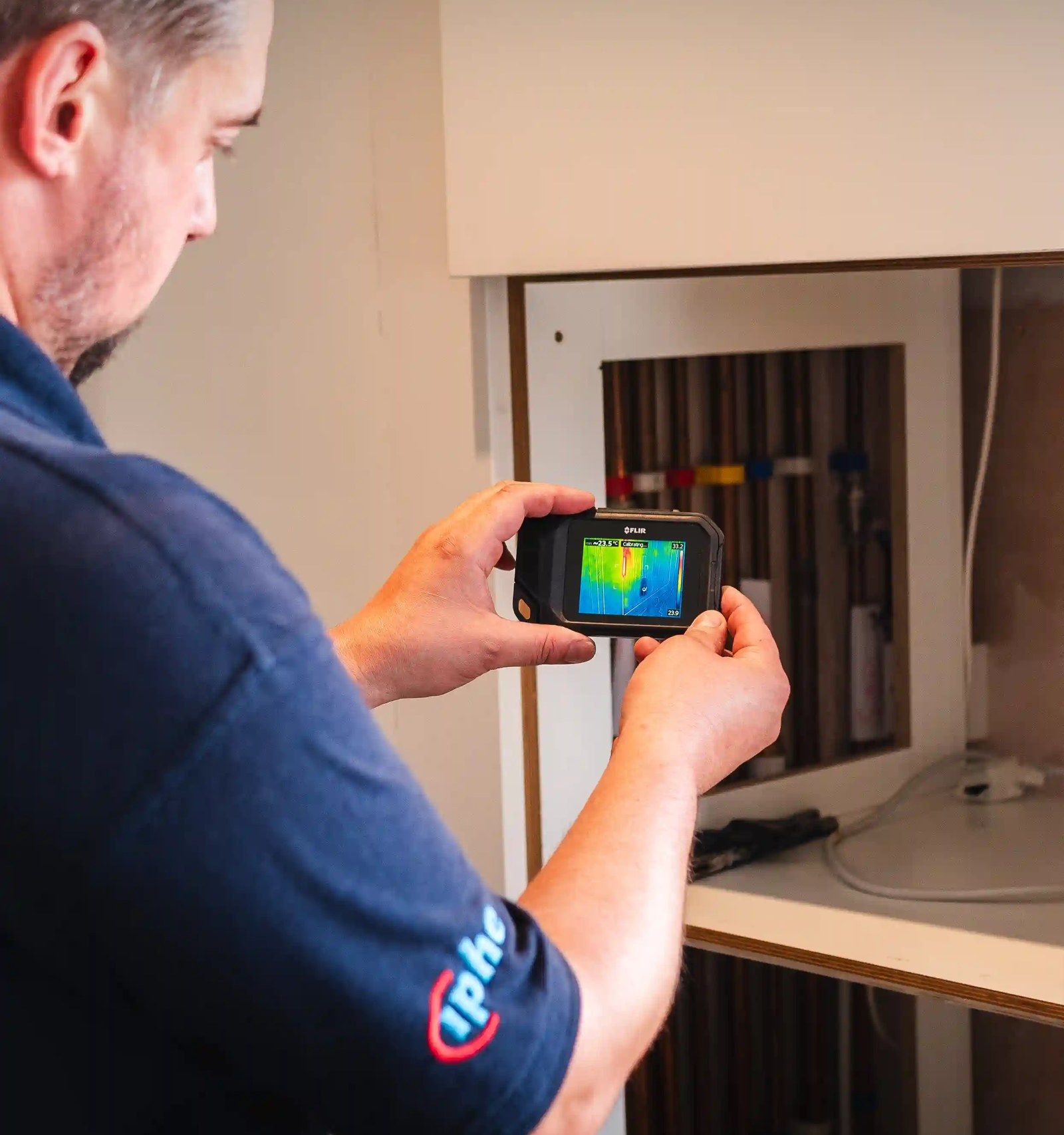 An engineer pointing a thermal imaging camera at an open cupboard of pipework
